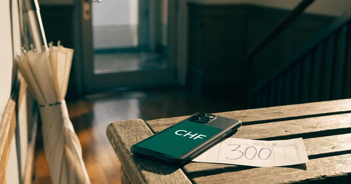 Ein Schlüsselbund und ein Smartphone mit dunklem Display liegen auf einer Holzbank in einem Hauseingang, daneben ein Regenschirm.