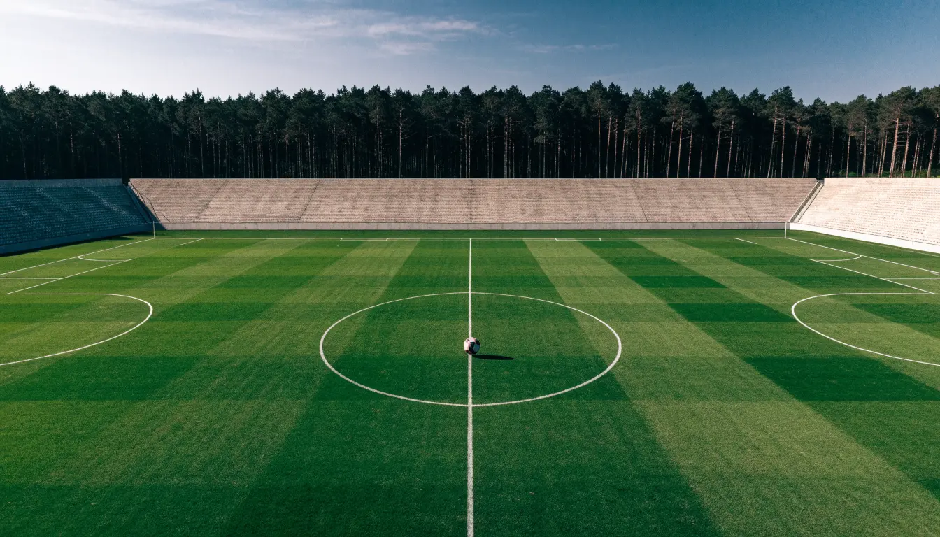 Ein Rasenplatz mit Spielfeldlinien ist von oben gesehen, ein einzelner Ball liegt in der Mitte, rundherum stehen leere Tribünen und Tannenwald.