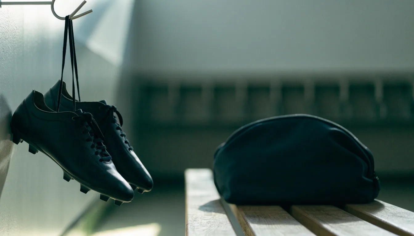 Ein Paar Fussballschuhe hängt an einem Haken in einer ruhigen Umkleide, darunter steht eine Sporttasche auf einer Holzbank.