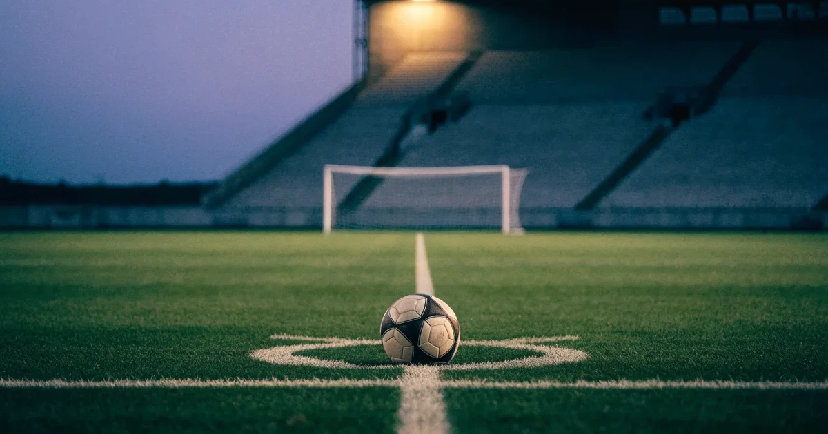 Ein Fussball liegt auf dem Mittelpunkt eines leeren Rasens, im Hintergrund steigen steile Tribünen in der Dämmerung auf.