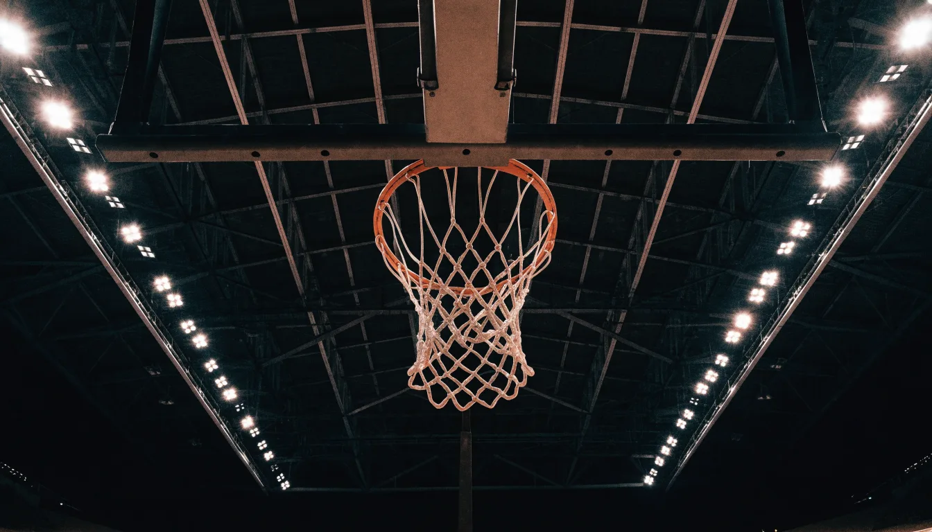 Ein Basketballkorb von unten gesehen, darüber eine Hallendecke mit Scheinwerfern und Stahlträgern.