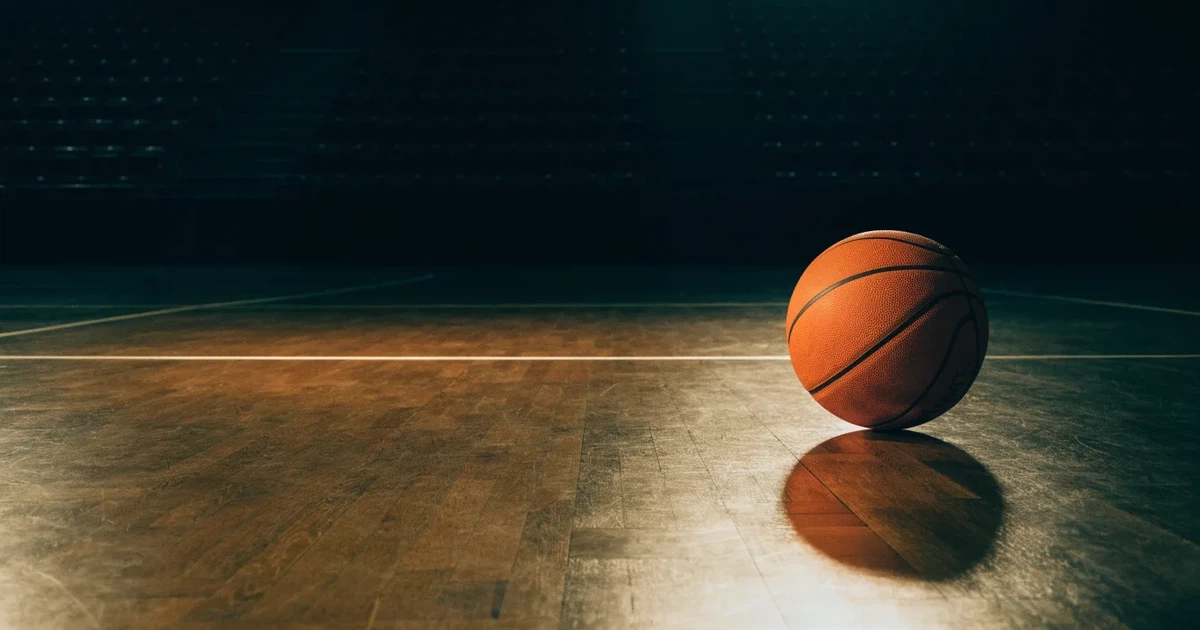 Ein orangefarbener Basketball liegt auf dem Parkett einer leeren Halle unter hellem Flutlicht, die Tribünen sind dunkel.