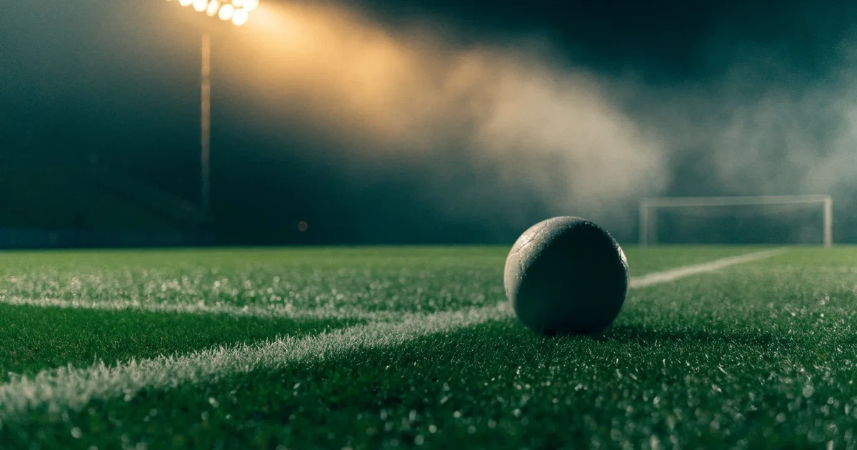 Ein Fussball liegt auf der Strafraumlinie eines nassen Rasens, dahinter stehen Flutlichtmasten im aufsteigenden Nebel.