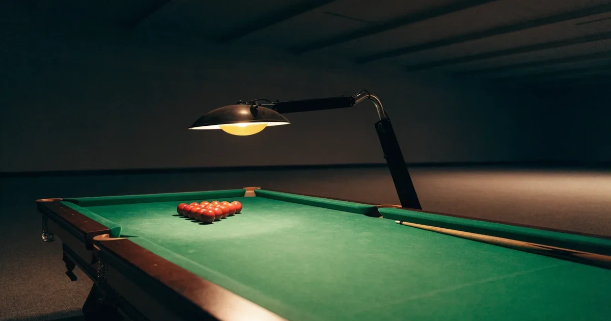 Ein Snookertisch mit grünem Tuch und einem Dreieck aus roten Kugeln unter einer tief hängenden Lampe in einer leeren Halle.