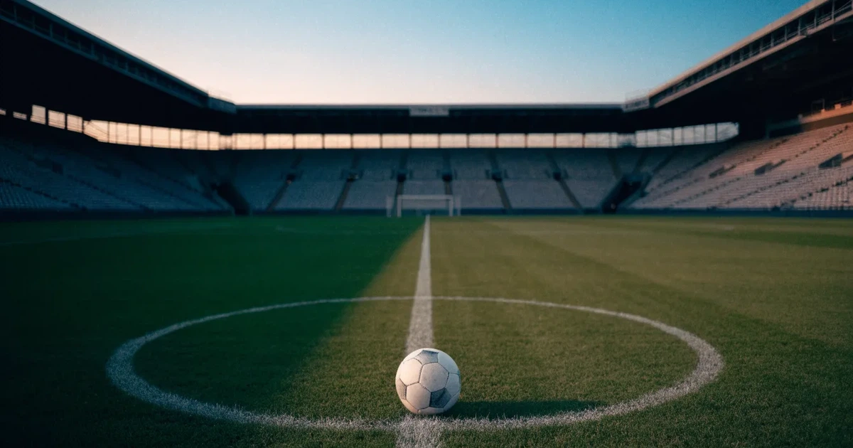 Ein Fussball liegt auf dem Anstosspunkt eines leeren Rasenplatzes, dahinter ragen die Ränge eines grossen Stadions in der Abenddämmerung auf.