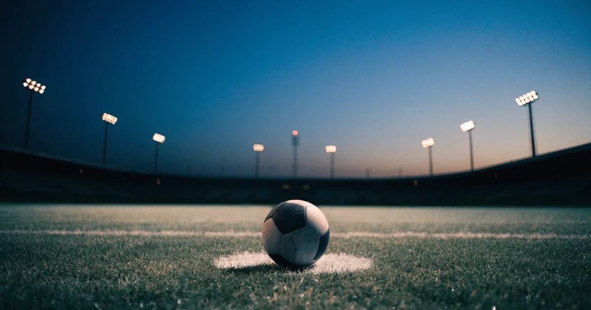 Ein Fussball liegt auf dem Anstosspunkt eines leeren Rasenplatzes, im Hintergrund glimmen Flutlichtmasten im Abendlicht.